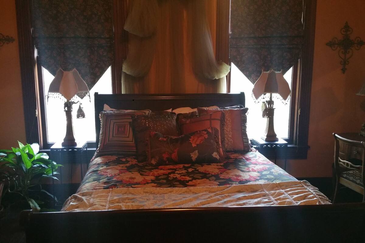Un dormitorio con una cama, dos ventanas con cortinas, un aparador y una lámpara.