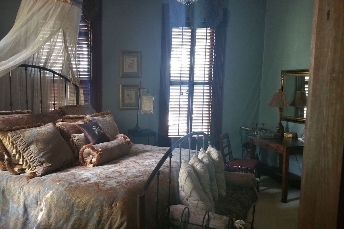 Un dormitorio con cama con dosel, una silla de madera, un escritorio con espejo y dos ventanas con persianas.