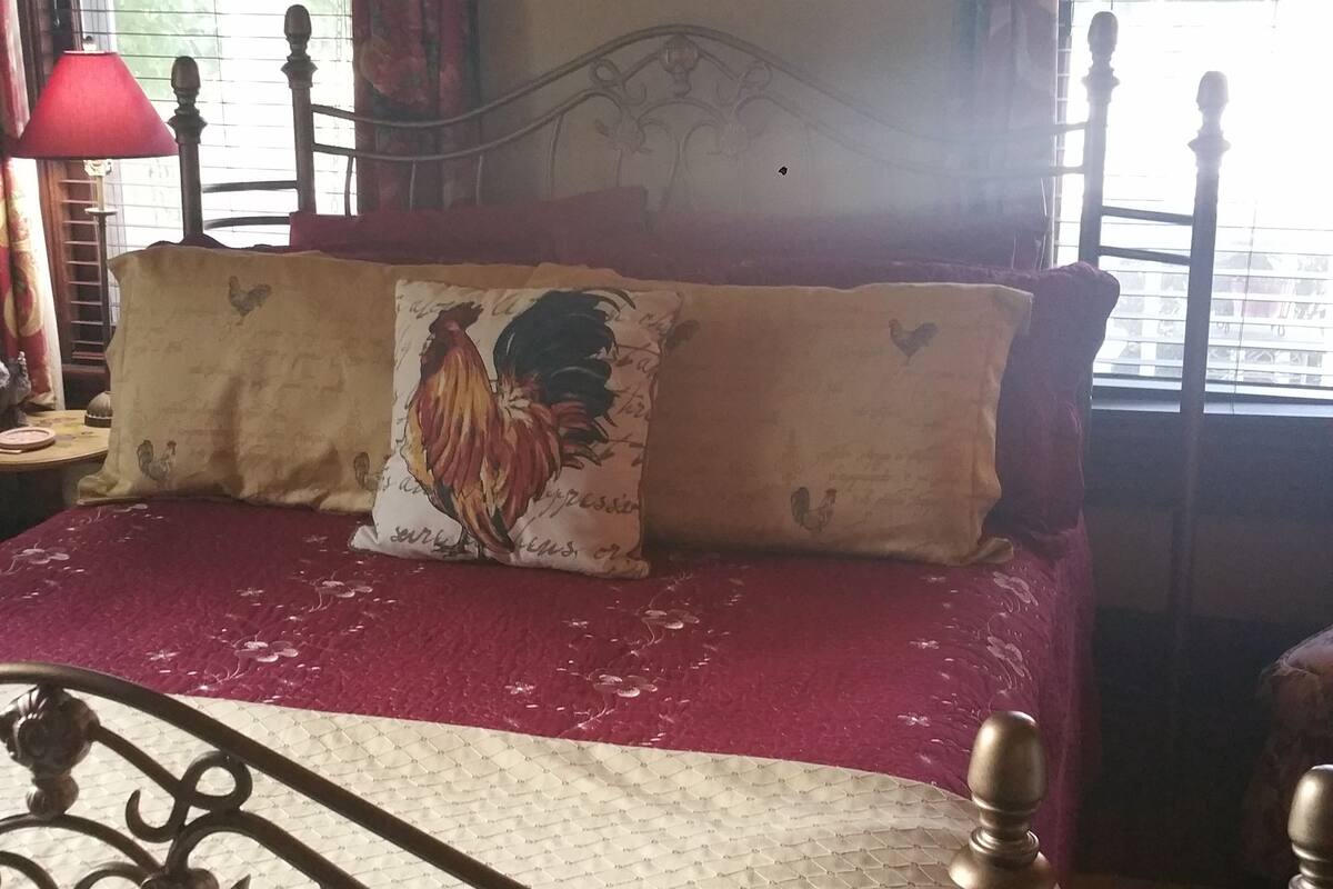 Una habitación con cama, una ventana con cortinas, una lámpara y un cuadro de gallos.