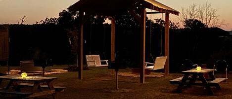 A wooden gazebo with picnic tables and string lights, surrounded by a grassy area and trees.