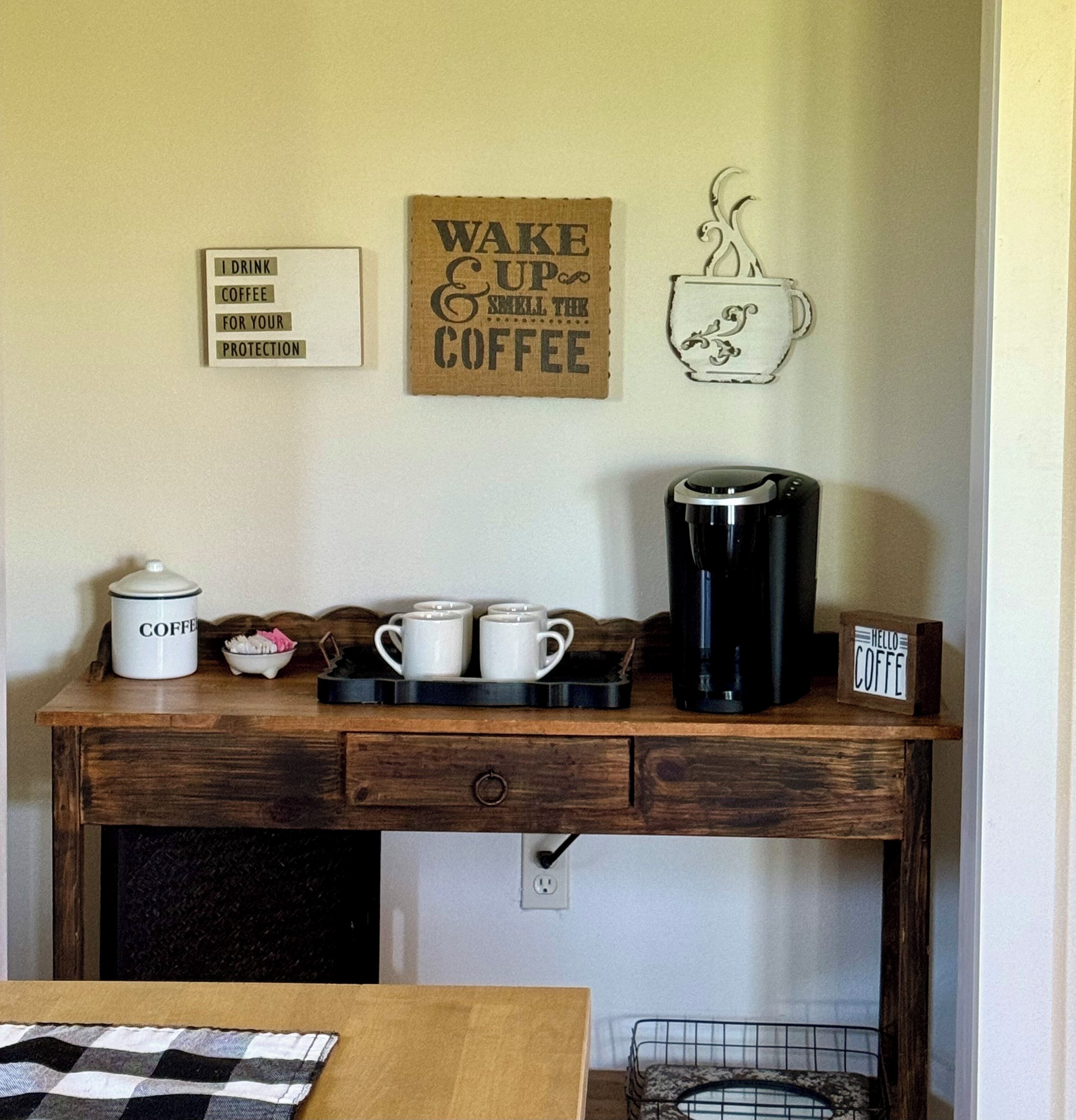 Una encimera de cocina con una cafetera, una jarra de café y una bandeja con tazas. Hay carteles en la pared que dicen “Bebo café por tu protección” y “Despierta con el aroma del café.”