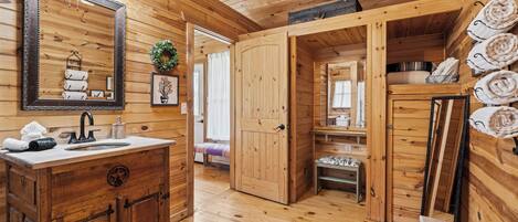 Interior de una cabaña de madera con tocador, espejo, lavamanos y una puerta que da a una zona de descanso.