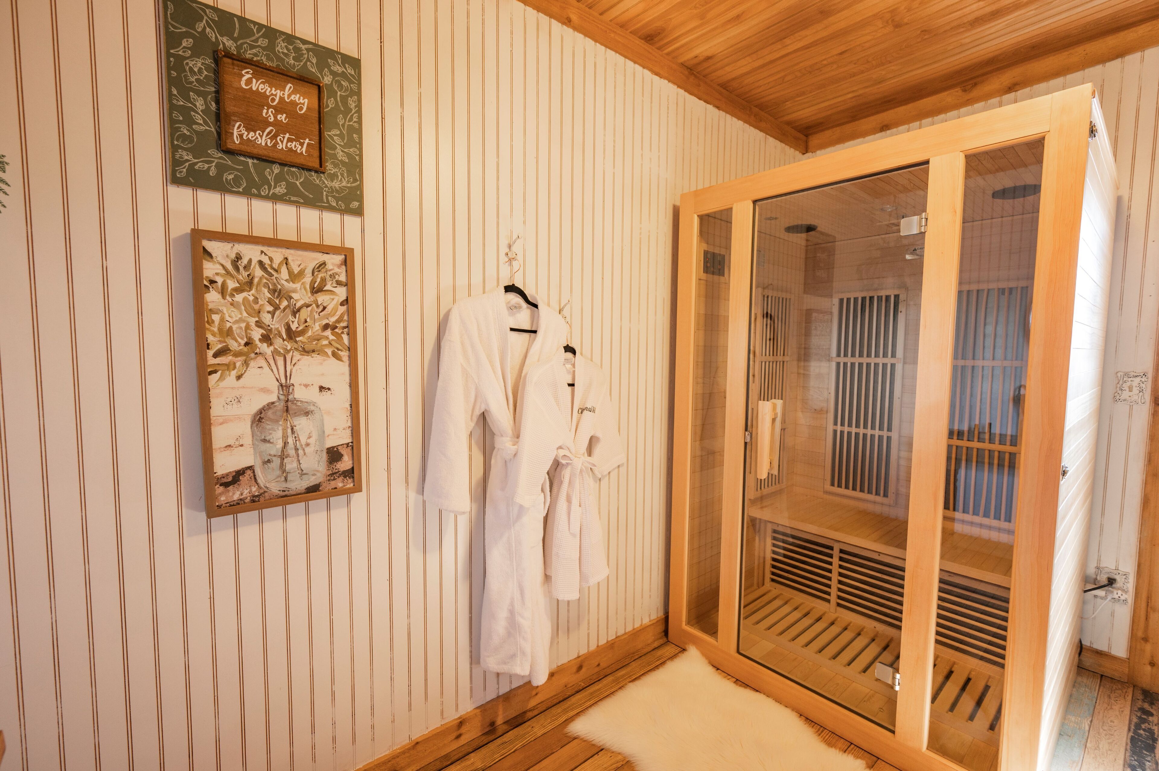 A room with wooden paneling, a glass door leading to a sauna, and two white robes hanging on the wall.