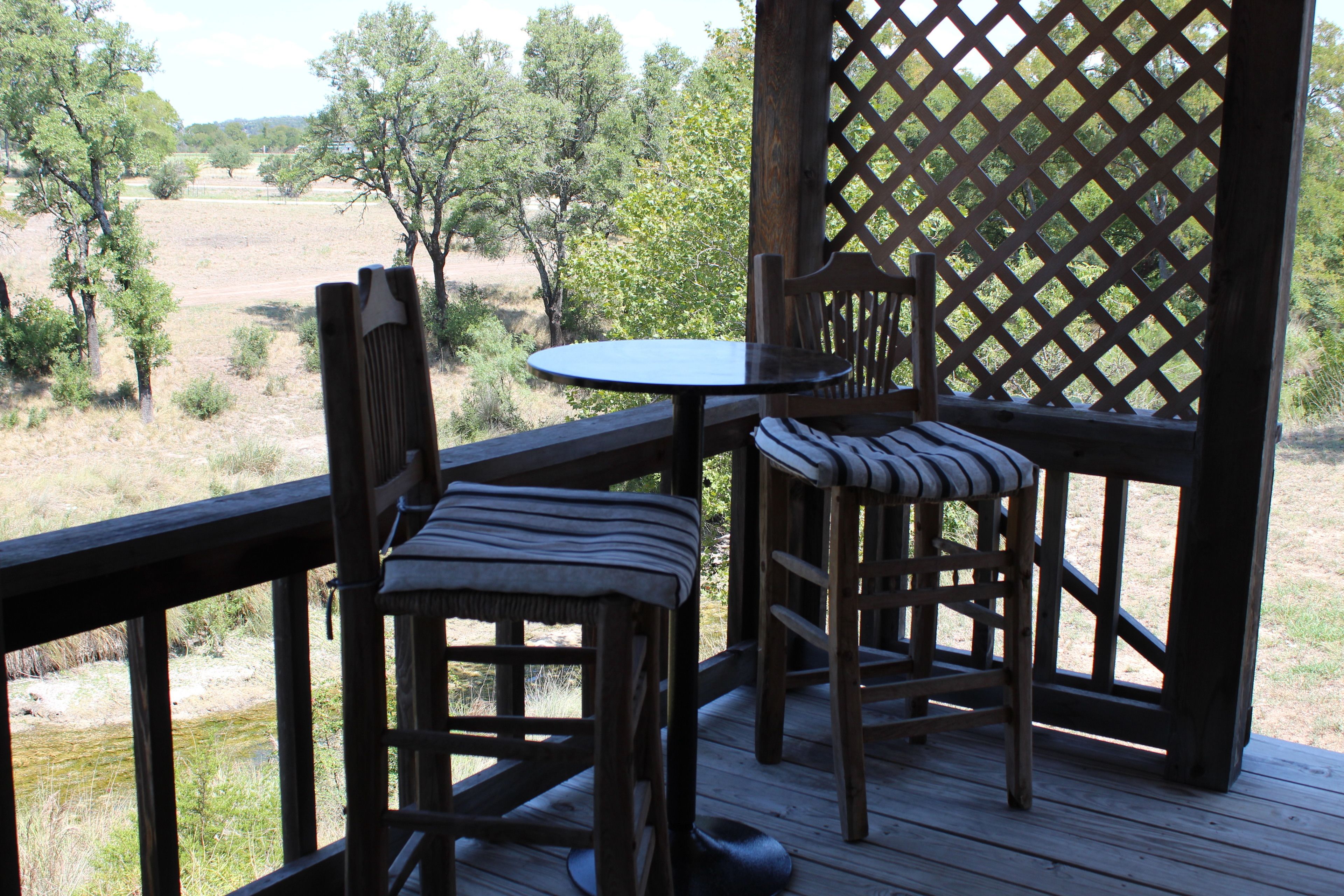 A wooden deck with a table and two chairs, overlooking a natural landscape with trees and a field.