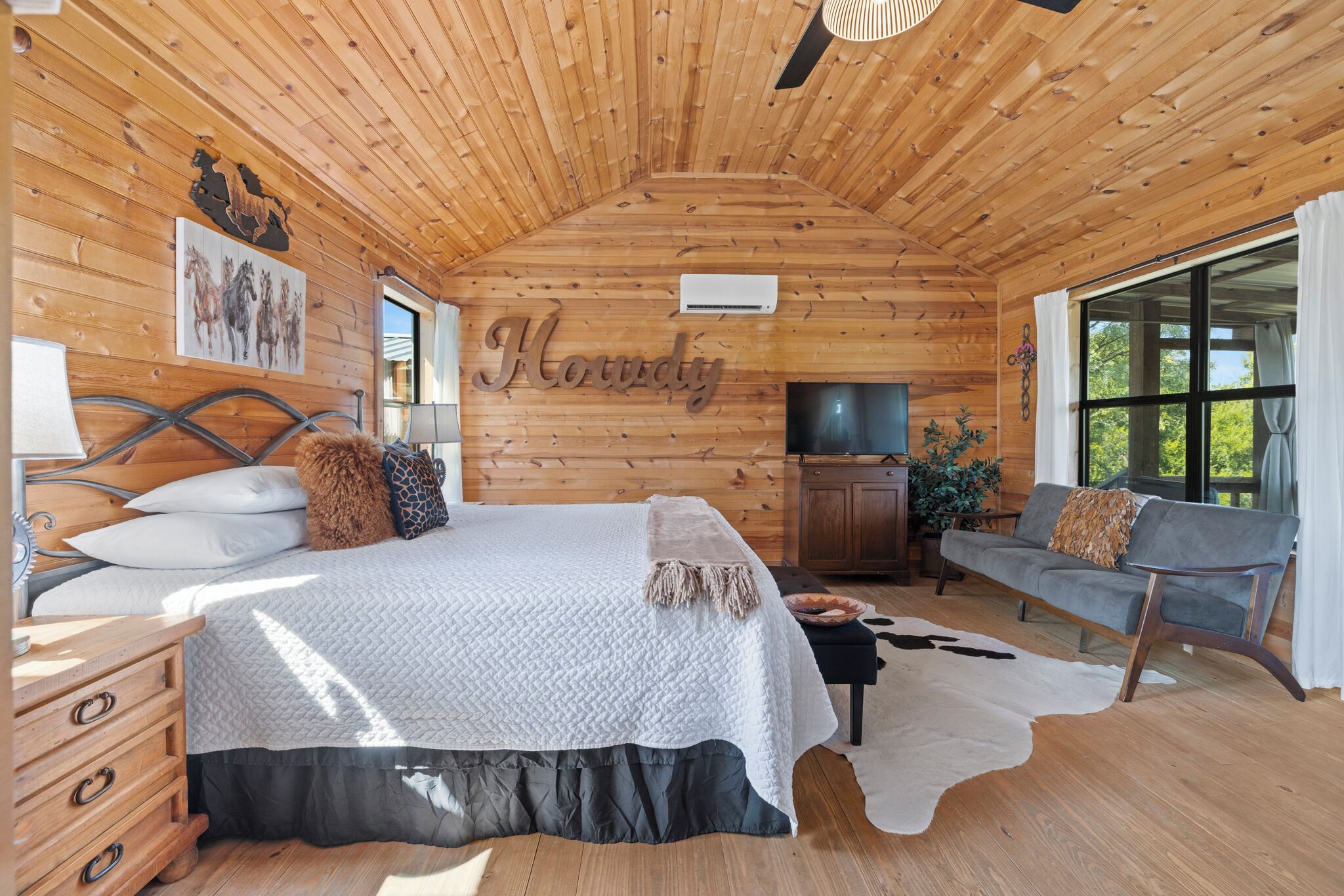 Un dormitorio de cabaña de madera con cama, sofá, televisor y un ventanal grande.