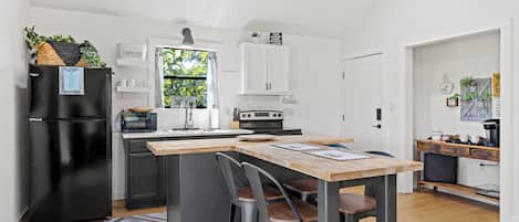Une cuisine moderne avec un réfrigérateur noir, un îlot central en bois doté de tabourets, des meubles blancs et une fenêtre donnant sur les arbres.