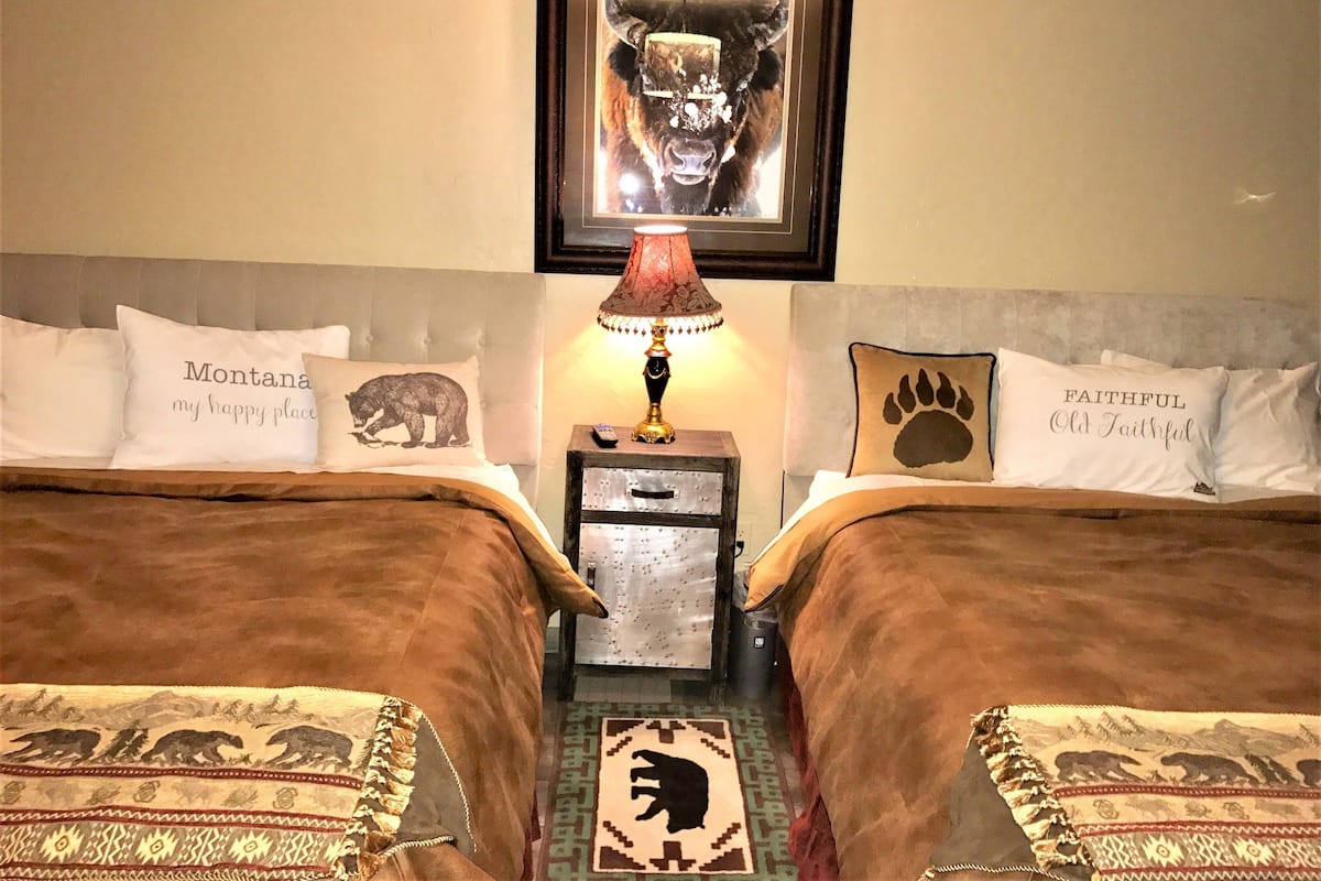 A hotel room with two beds, each with a bear-themed pillow, a framed picture of a bear on the wall, and a small nightstand with a lamp.