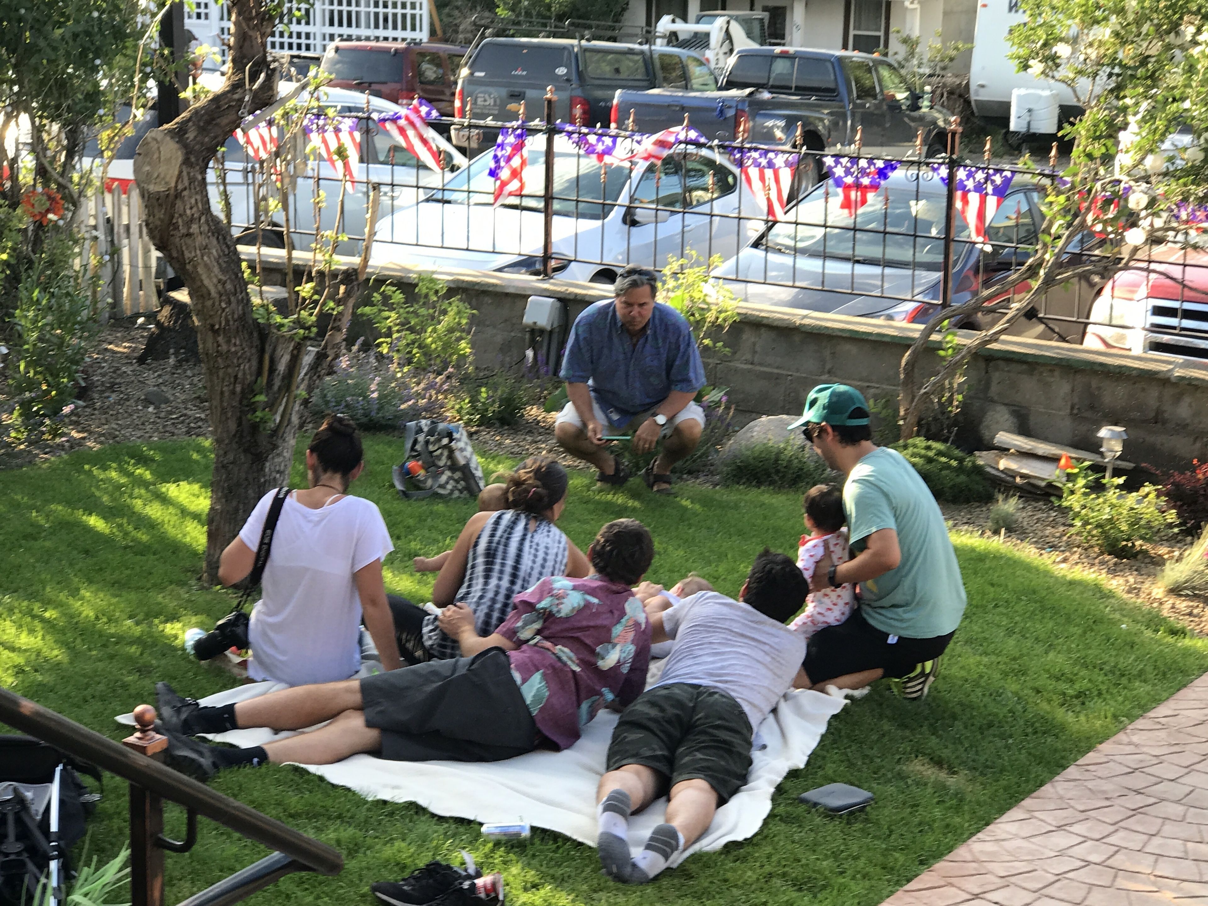 A group of people sitting on a blanket in a grassy area with a metal fence and parked cars in the background.