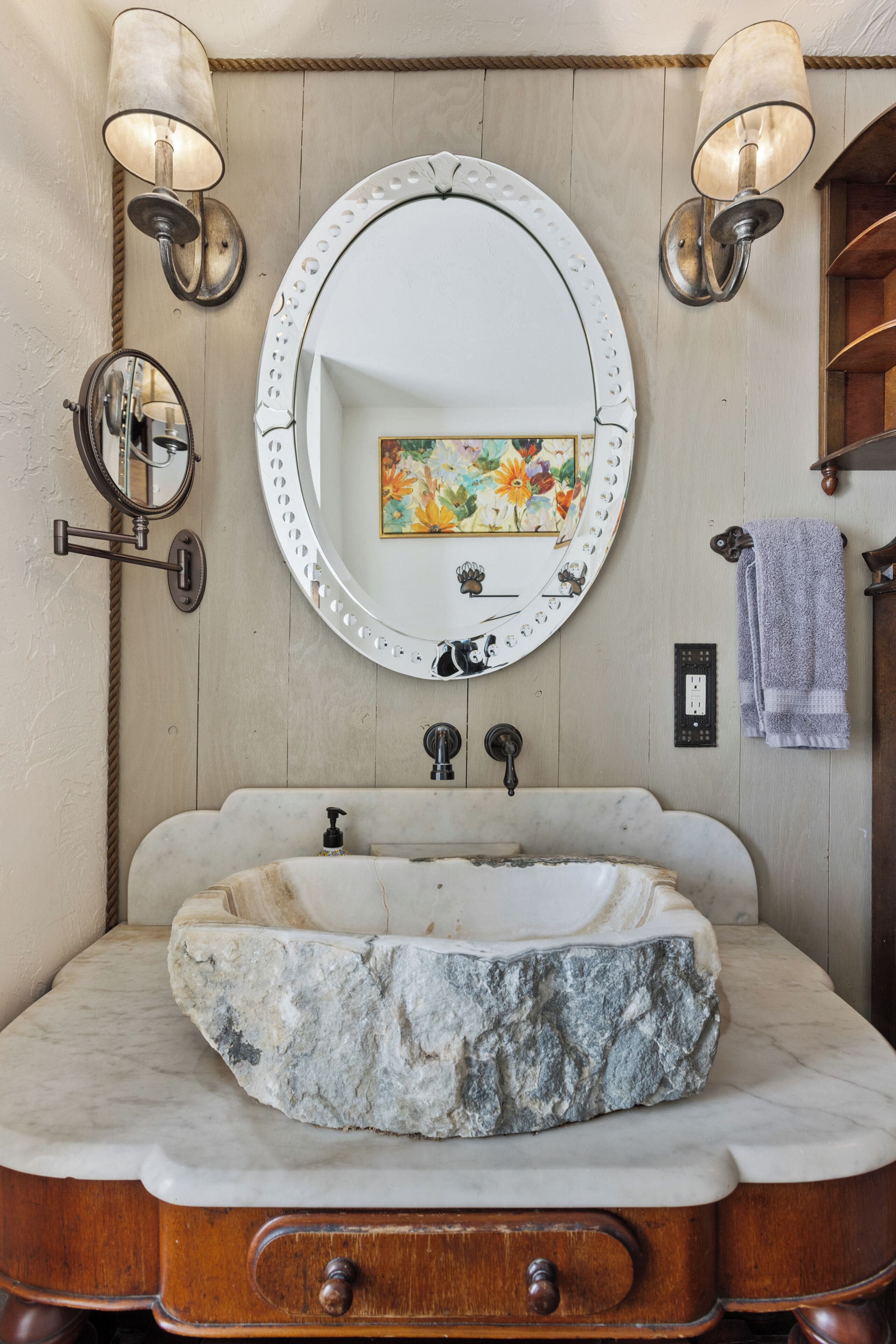 A bathroom with a marble sink, wooden vanity, two wall-mounted lights, a round mirror, and a towel rack.