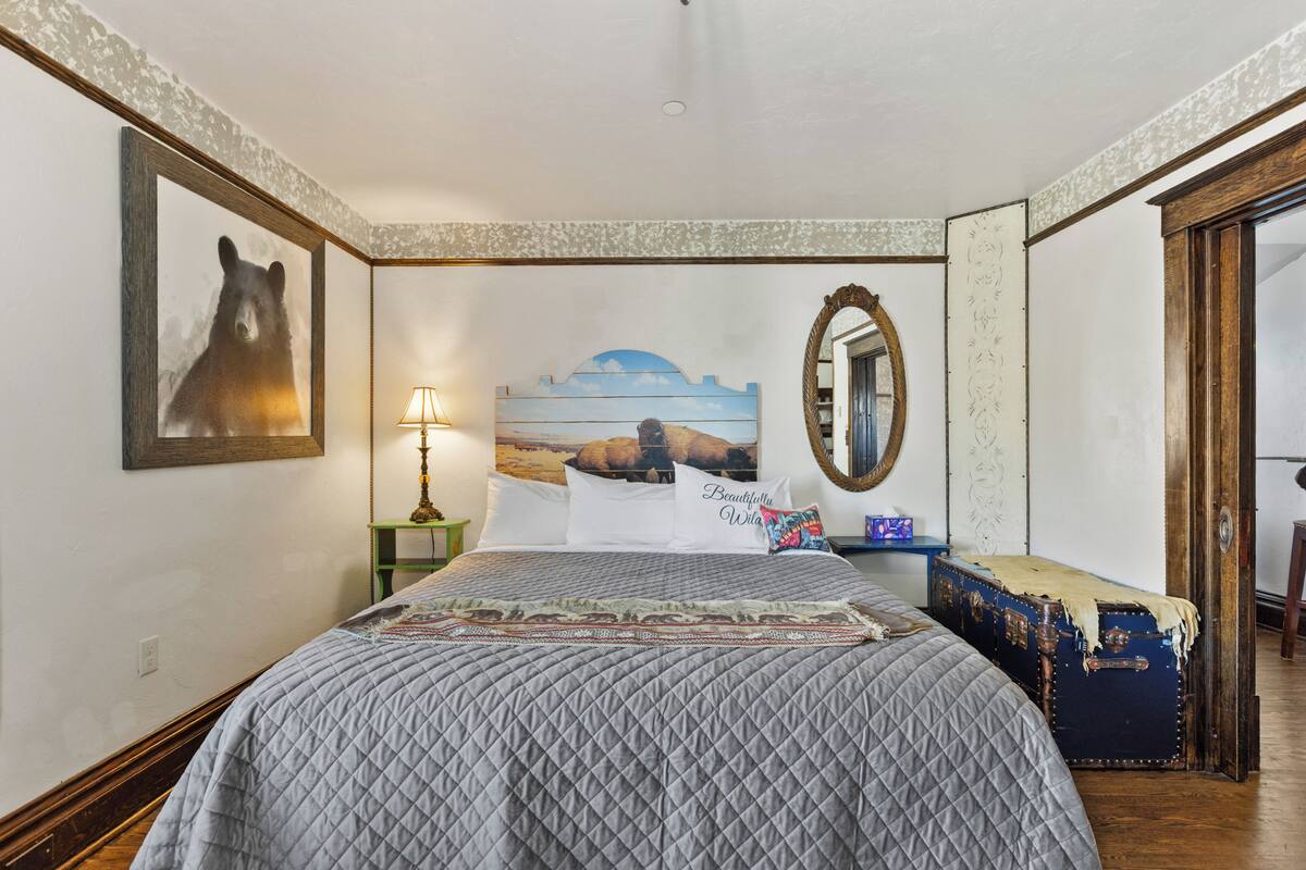 A bedroom with a quilted bedspread, a framed bear picture, a landscape painting, a lamp, and a vintage suitcase.