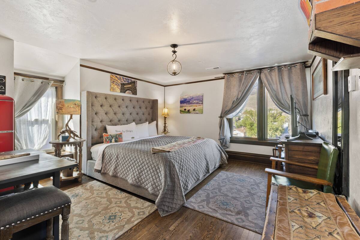 A bedroom with a large bed, a desk, a chair, and a window with curtains.