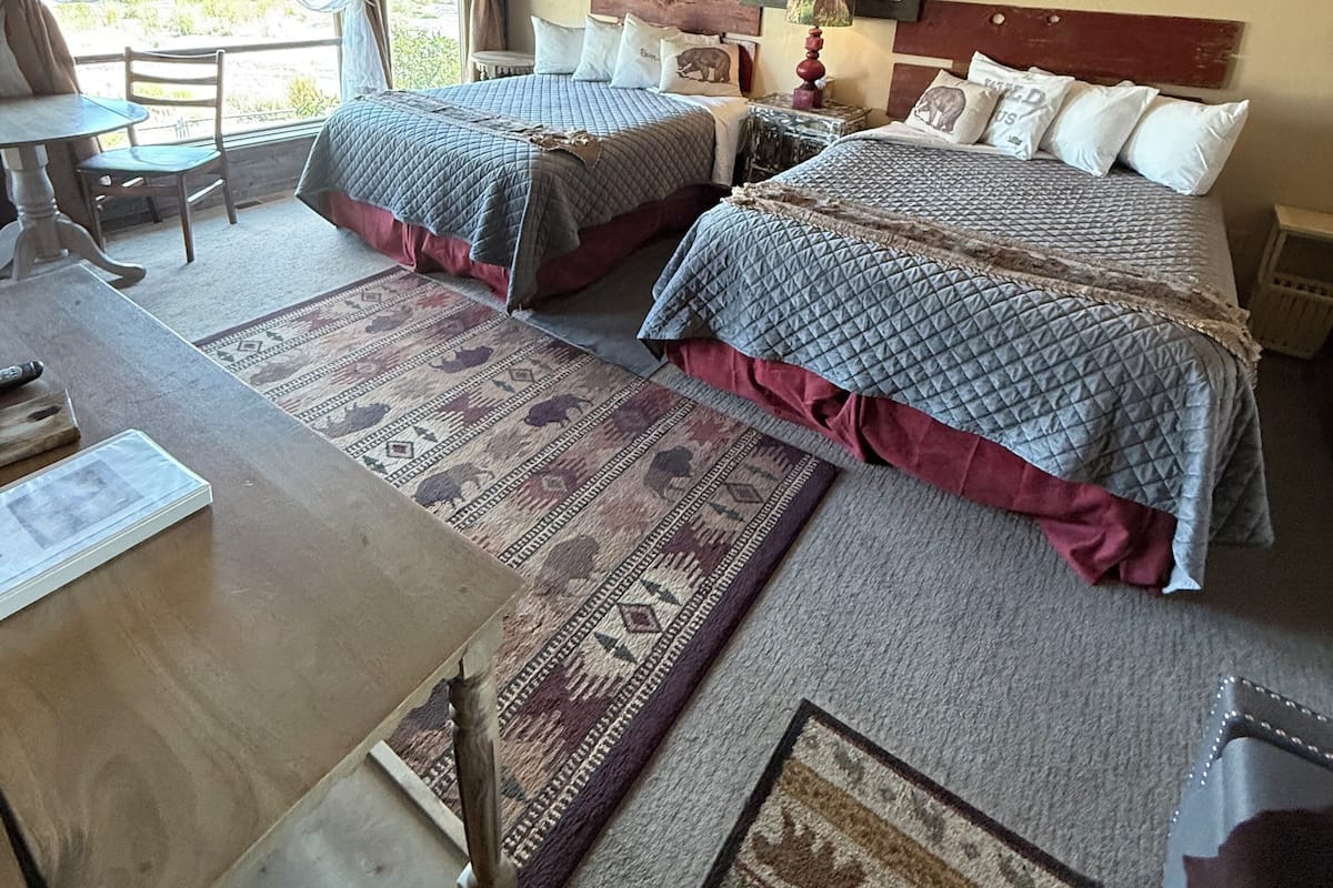 A hotel room with two beds, a dining table, a chair, a rug, and a bear-themed wall decoration.