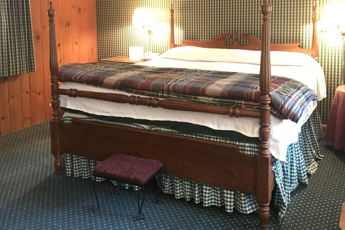 A four-poster bed with a plaid bedspread, a wooden headboard, and a footboard. There are two bedside lamps and a small table with a chair. The room has wooden walls and a patterned carpet.