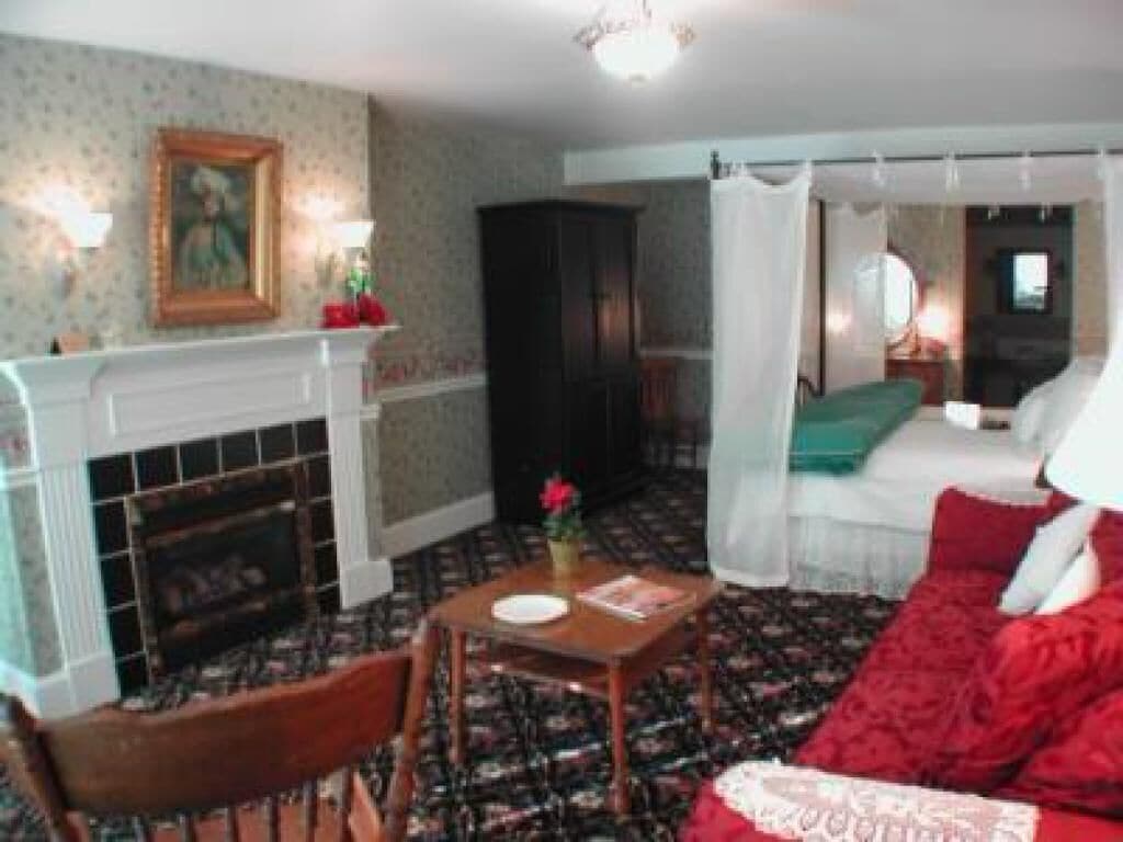 A room with a fireplace, a bed with red bedding, a wooden chair, and a table with a vase of flowers.