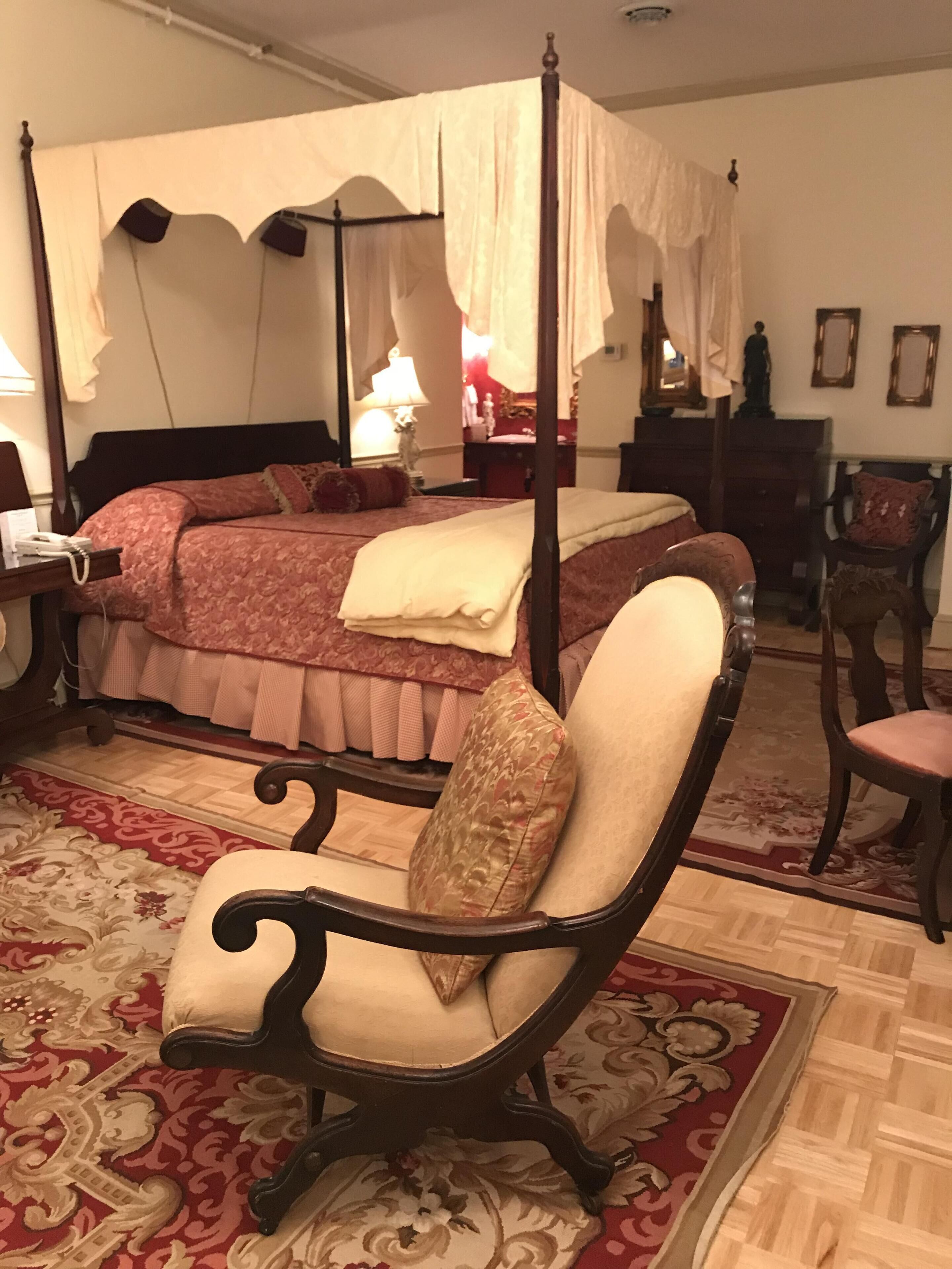 Una cama con dosel y cuatro columnas, con un edredón rojo y beige, una silla de madera con cojín y una alfombra estampada.