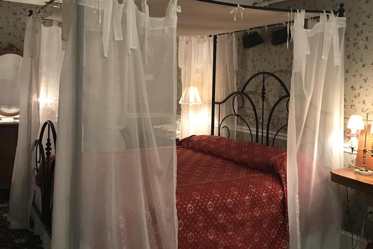 A four-poster canopy bed with a red patterned bedspread, a bedside table with a lamp, and a patterned carpet.