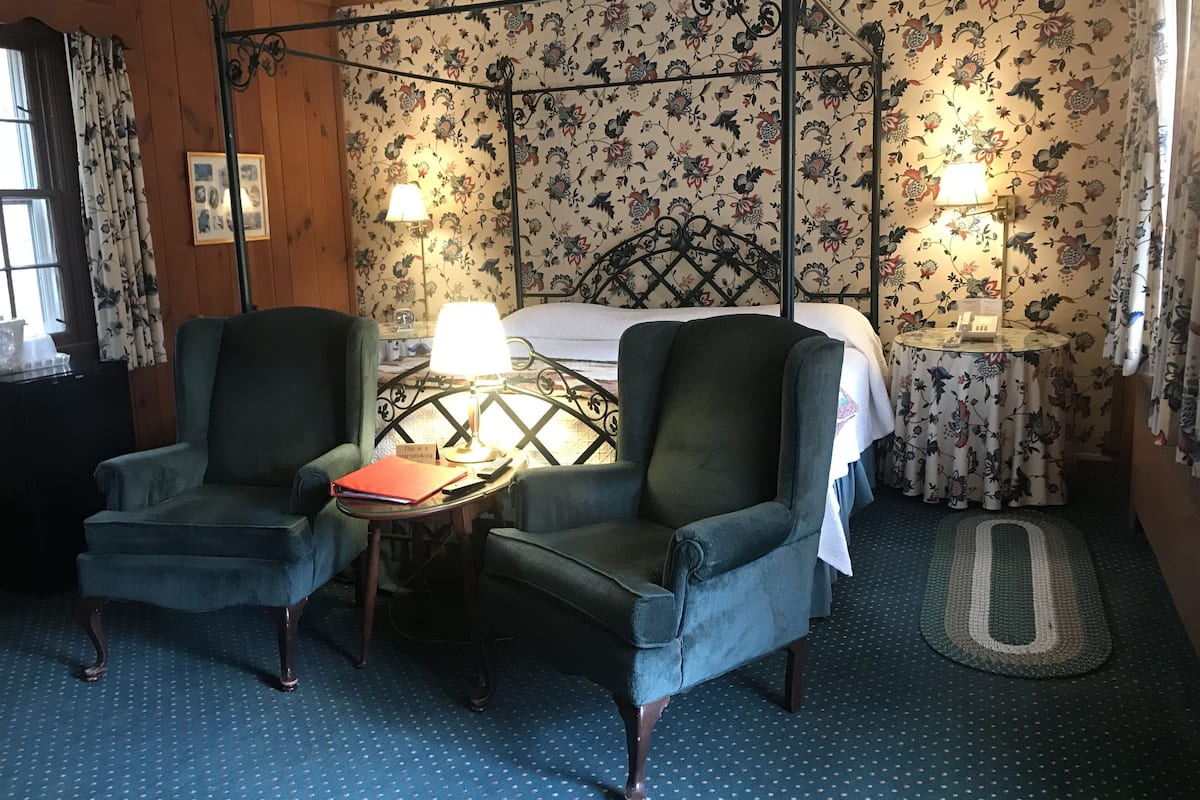 A room with two armchairs, a bed with floral wallpaper, two bedside lamps, and a small table with a red folder.