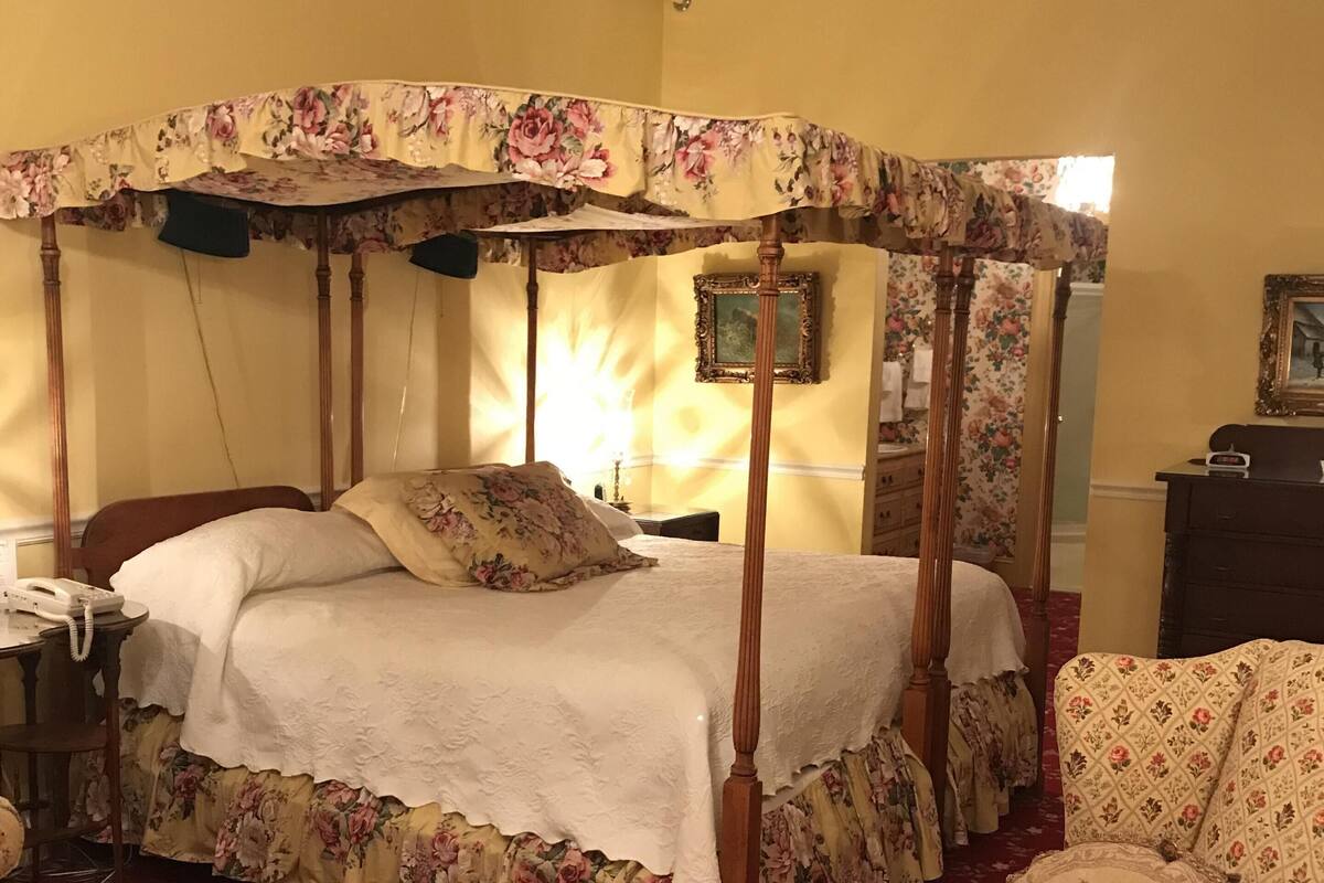 A four-poster canopy bed with floral upholstery, a wooden nightstand with a telephone, and a floral-patterned armchair.