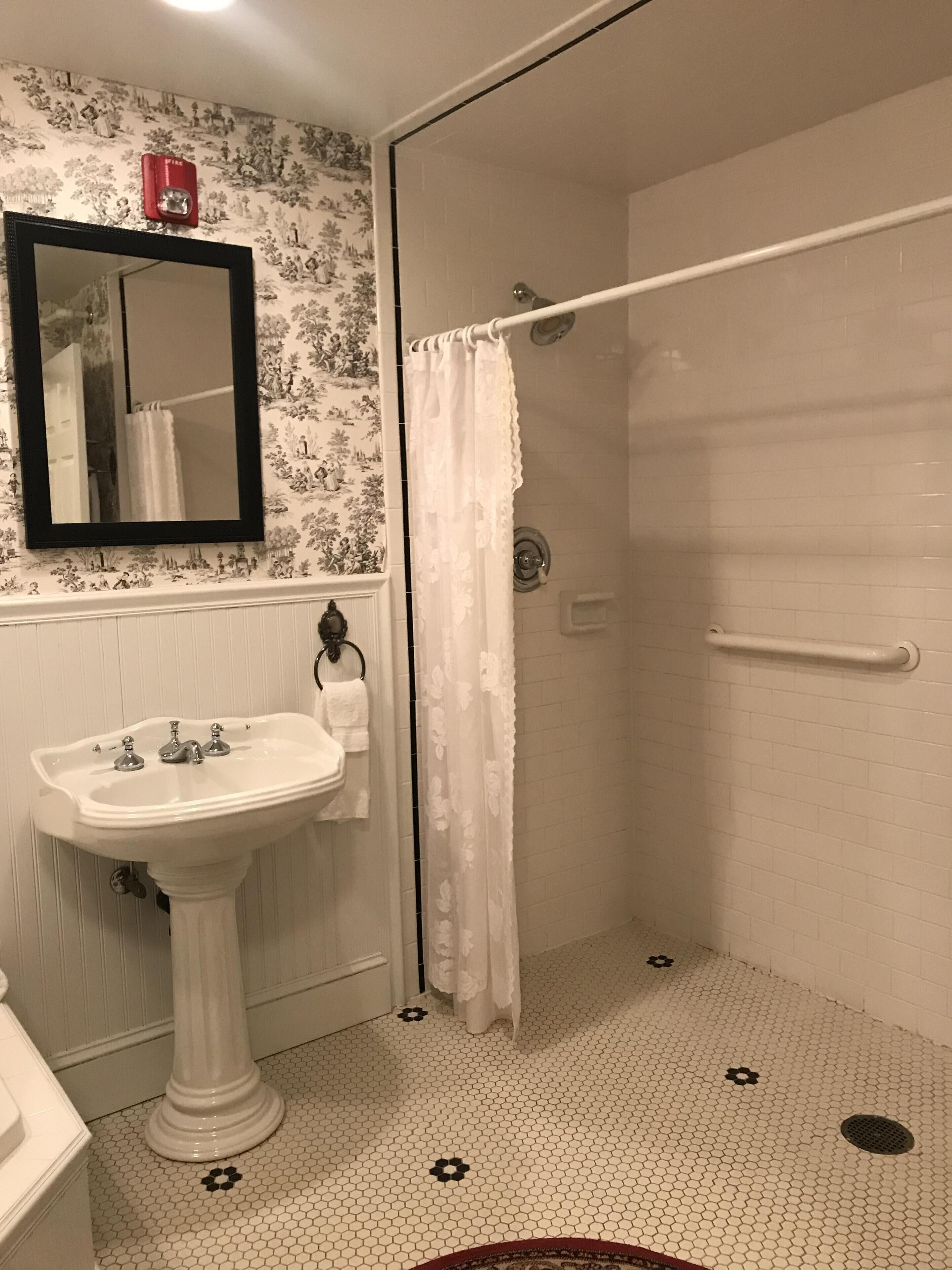 Un baño con un lavamanos blanco, un espejo, una ducha con cortina blanca y un suelo de baldosas con motivos florales negros.