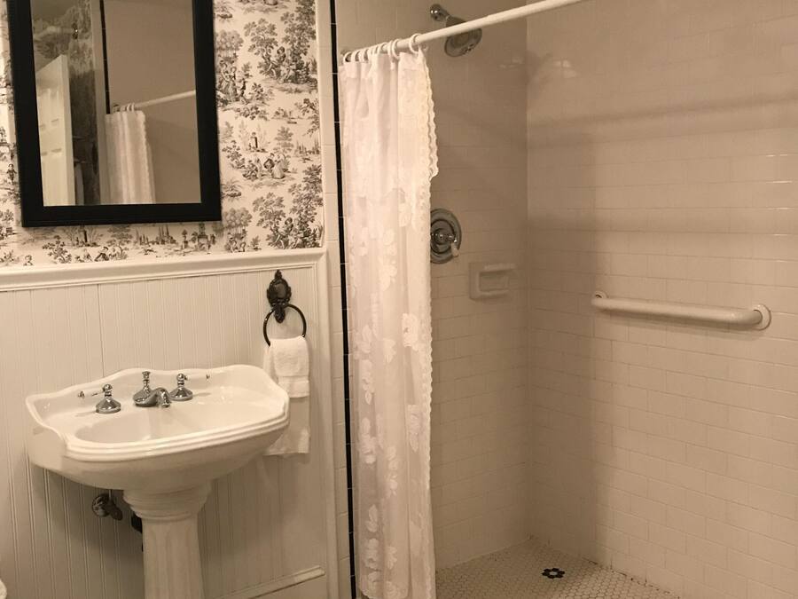 Un baño con un lavamanos blanco, un espejo, una ducha con cortina blanca y un suelo de baldosas con motivos florales negros.