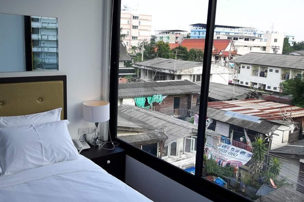 A hotel room with a bed, a bedside table with a lamp, and a view of the cityscape through large windows.