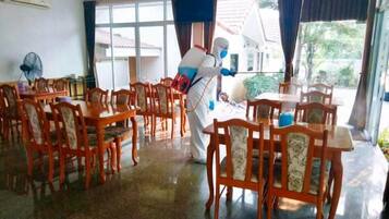 Un comedor con mesas y sillas de madera, ventanas amplias con cortinas, y un equipo de aire acondicionado en la pared.