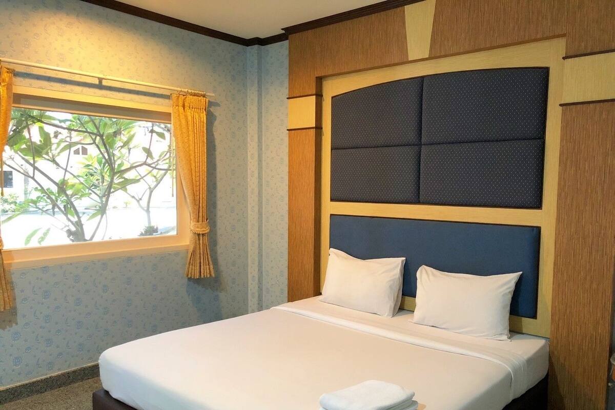 A neatly made bed with white linens and pillows, a window with curtains, and a headboard with a dark panel.