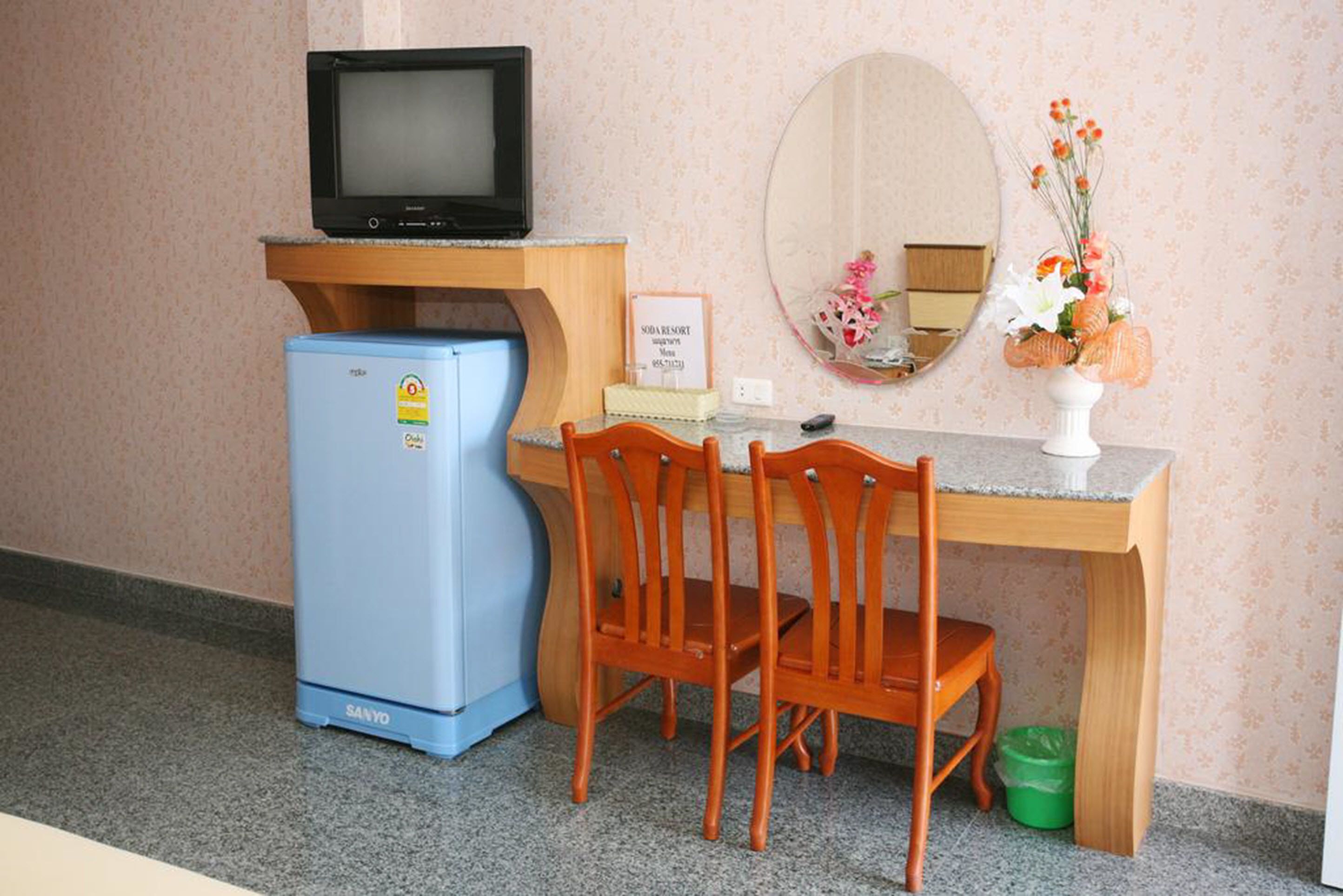 Une pièce de taille modeste, avec un petit réfrigérateur bleu, un bureau en bois sur lequel est posée une télévision, un miroir et un vase rempli de fleurs.