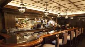 Uma área de bar com balcão polido, vários bancos altos e um bar bem abastecido com diversas garrafas e copos.