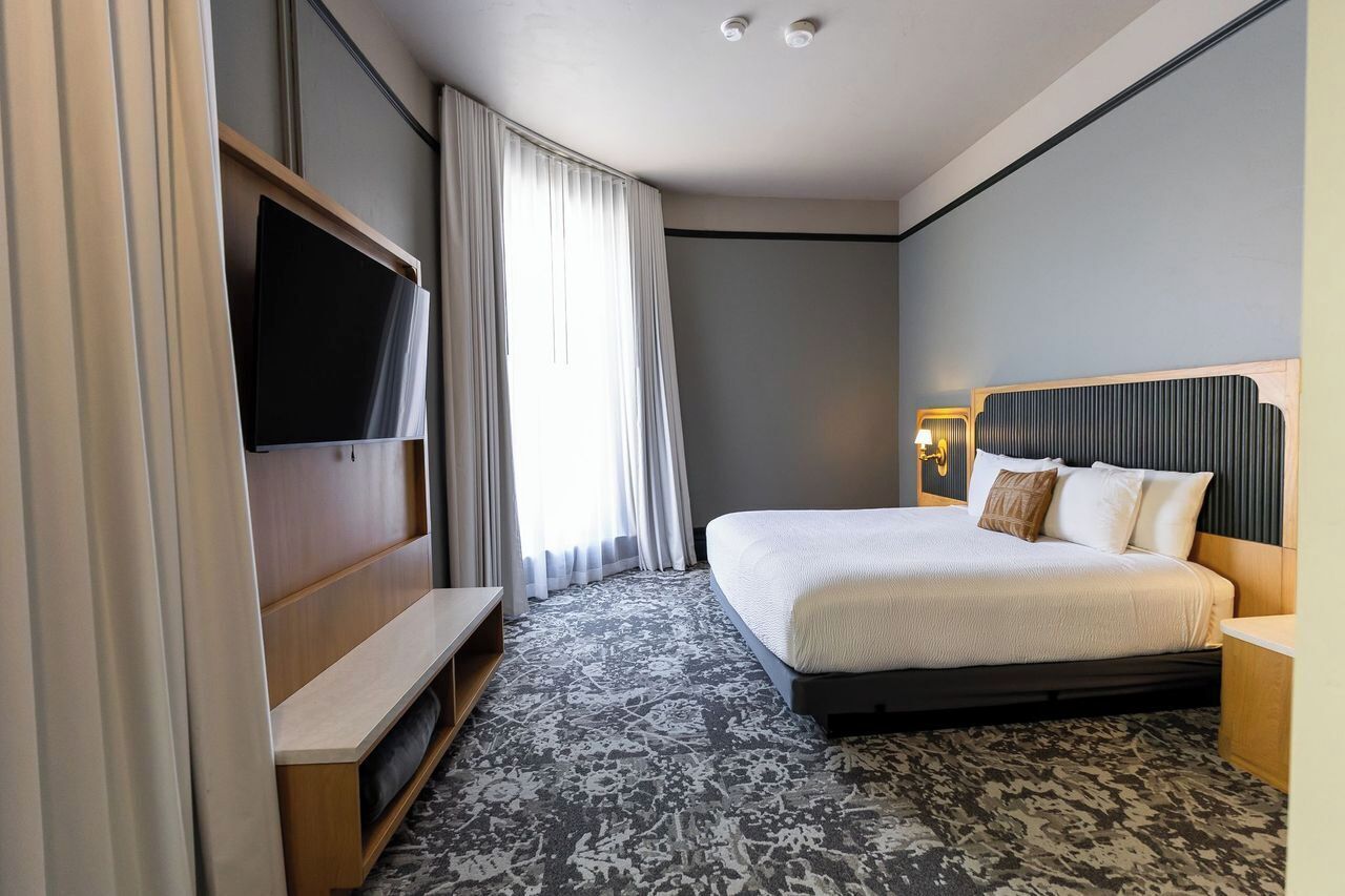 A hotel room with a bed, a TV, a desk, and a window with curtains.