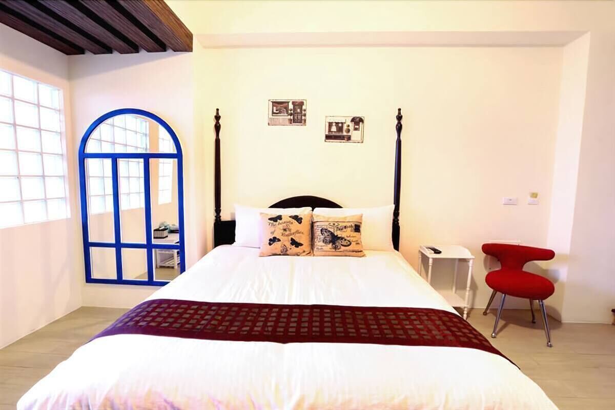 A bedroom with a bed, a red chair, a small table, and a window with a blue frame.