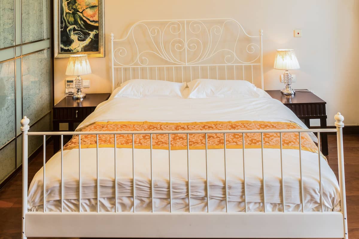 Una cama bien hecha con estructura metálica blanca, dos mesitas de noche con lámparas, un cuadro en la pared y un candelabro en el techo.