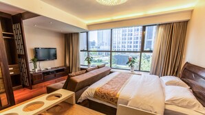 A hotel room with a large bed, a TV mounted on the wall, a seating area with a table, and a view of the city through large windows.
