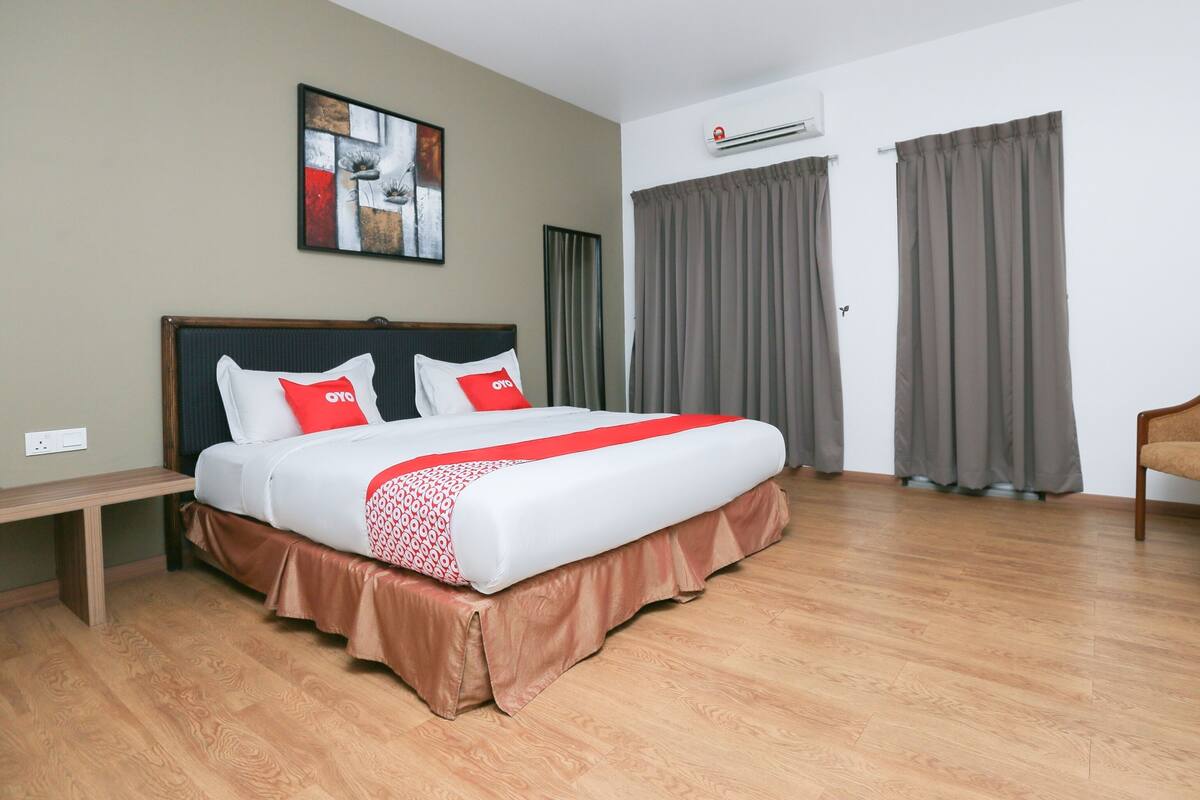 A hotel room with a large bed, a headboard, a bedside table, a wall-mounted air conditioner, and a window with curtains.
