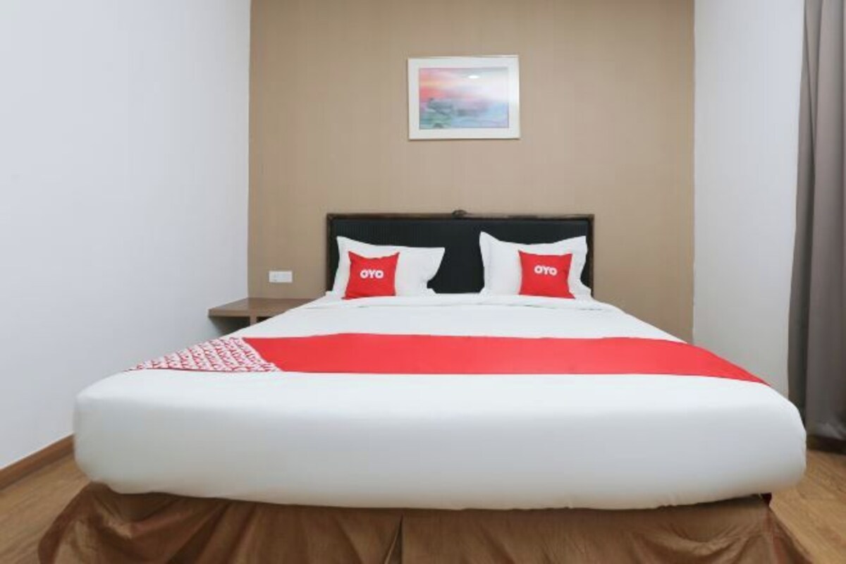 Un lit recouvert d’une couvre-lit blanche et rouge, deux oreillers ornés d’un logo rouge, une table de chevet et un tableau accroché au mur.