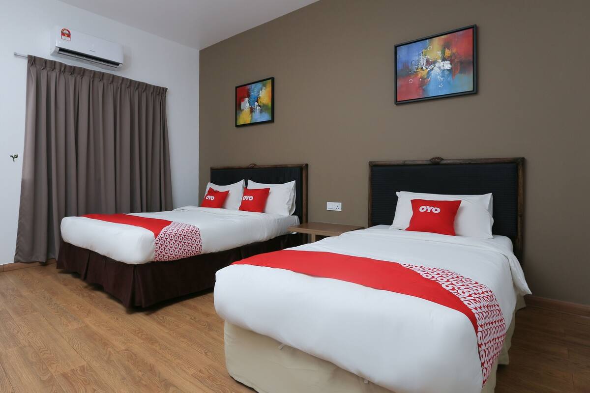 A hotel room with two beds, each with a red and white bedspread and matching pillows. There is a painting on the wall and an air conditioning unit above the window.