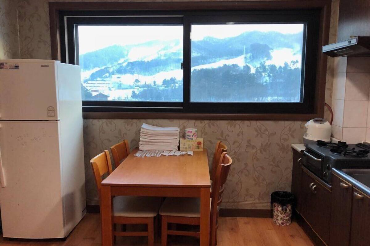 A kitchen with a wooden table, chairs, a refrigerator, a stove, and a window with a view of a snowy landscape.