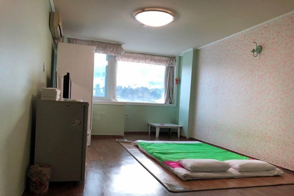 A small, clean room with a bed, a small table, a window, and a refrigerator.