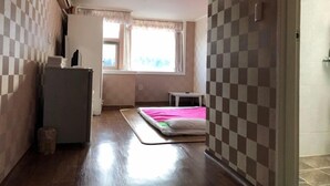 Una habitación pequeña y funcional, con cama, una mesita y un refrigerador. Tiene una ventana con cortinas y vistas al exterior.