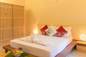 A neatly made bed with white linens, two red decorative pillows, and a pair of white swan-shaped towels. A wooden nightstand with a lamp is beside the bed. - Thoddoo Retreat (Thoddoo)