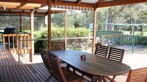 Una terrassa de fusta a l'exterior amb taula rodona i cadires, una barbacoa i una zona coberta amb vistes a vegetació.