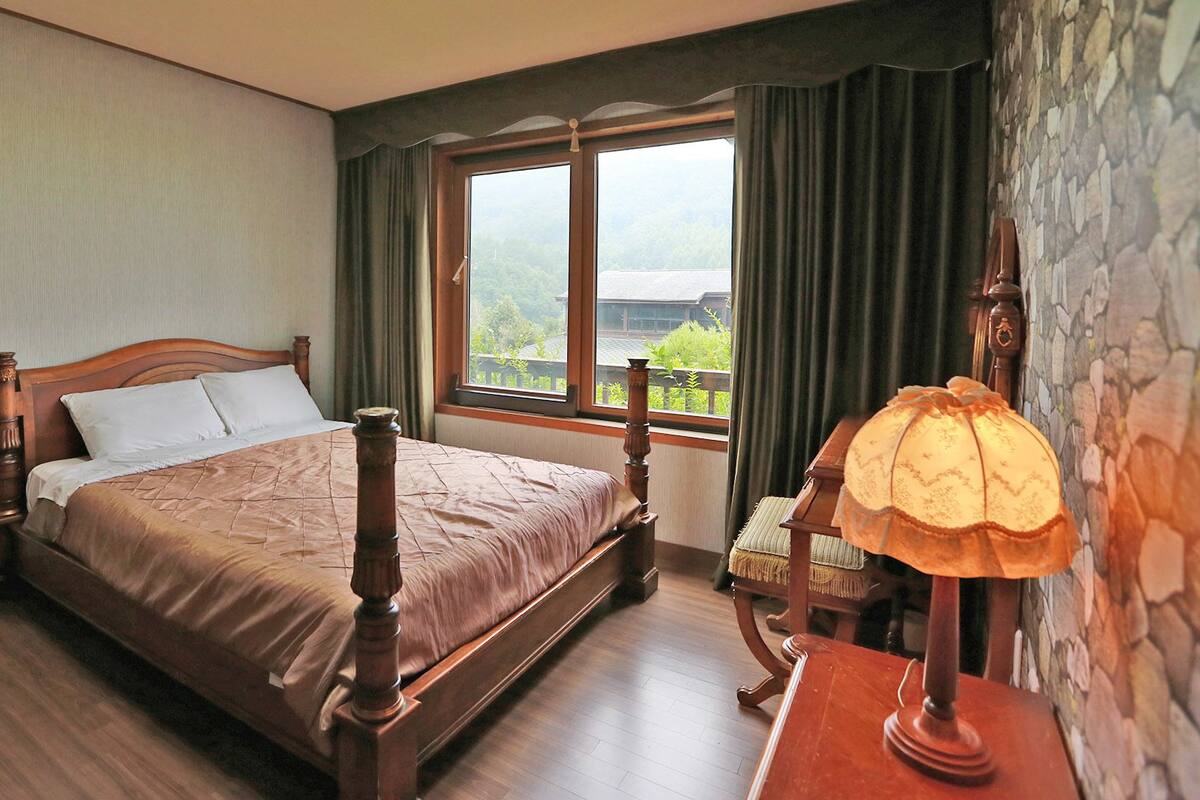 A neatly made bed with a wooden headboard, a window with a view of greenery, and a lamp on a wooden nightstand.