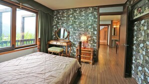 A bedroom with a bed, a dresser, a mirror, and a lamp. There is a stone wall and a wooden door leading to another room. - Chalet Resort (Pyeongchang)