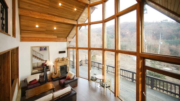 A spacious living room with a fireplace, large windows, and a view of a deck and surrounding landscape. - Chalet Resort (Pyeongchang)