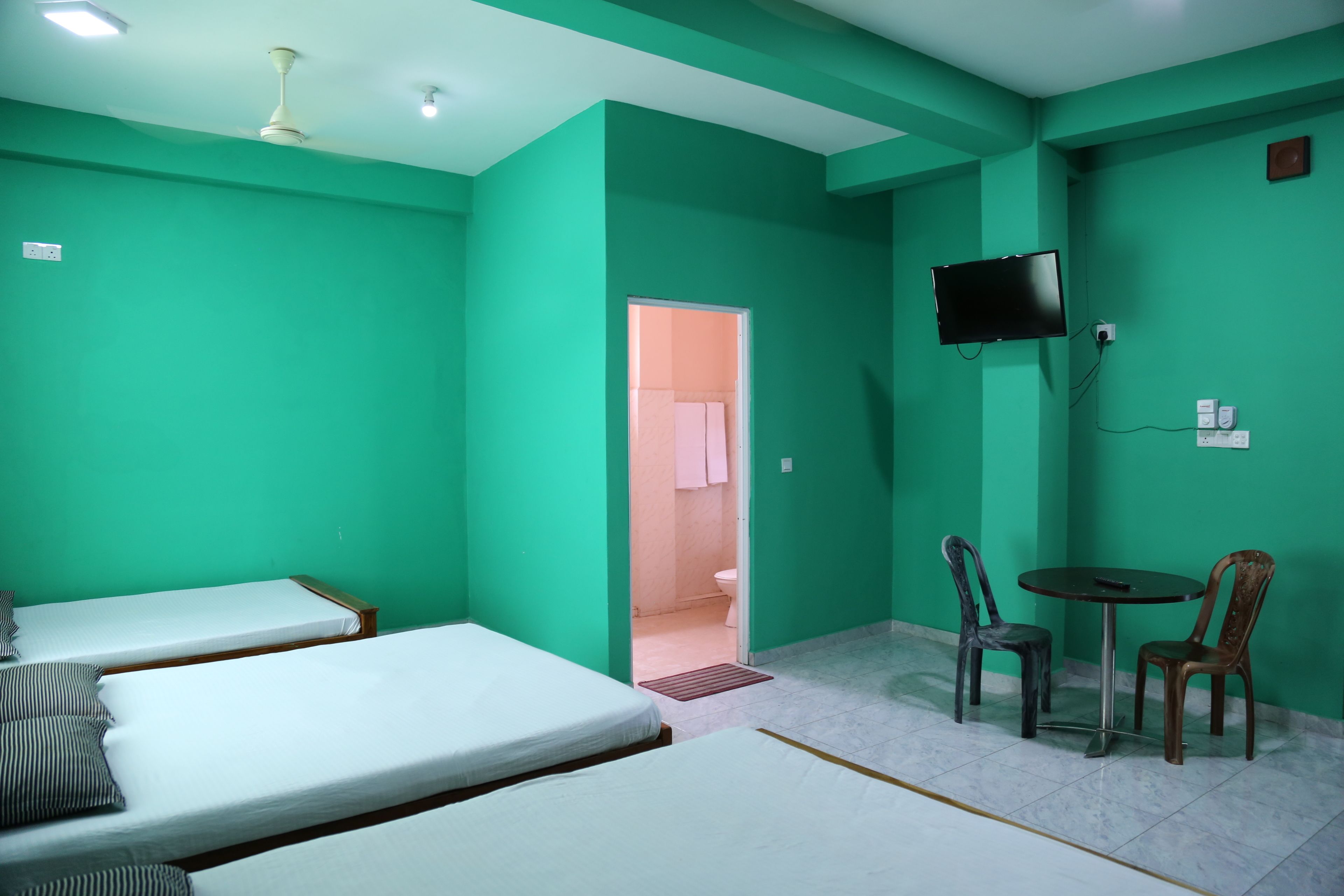 Une chambre avec deux lits simples, une petite table avec deux chaises, un téléviseur fixé au mur et une salle de bain visible à travers une porte ouverte.