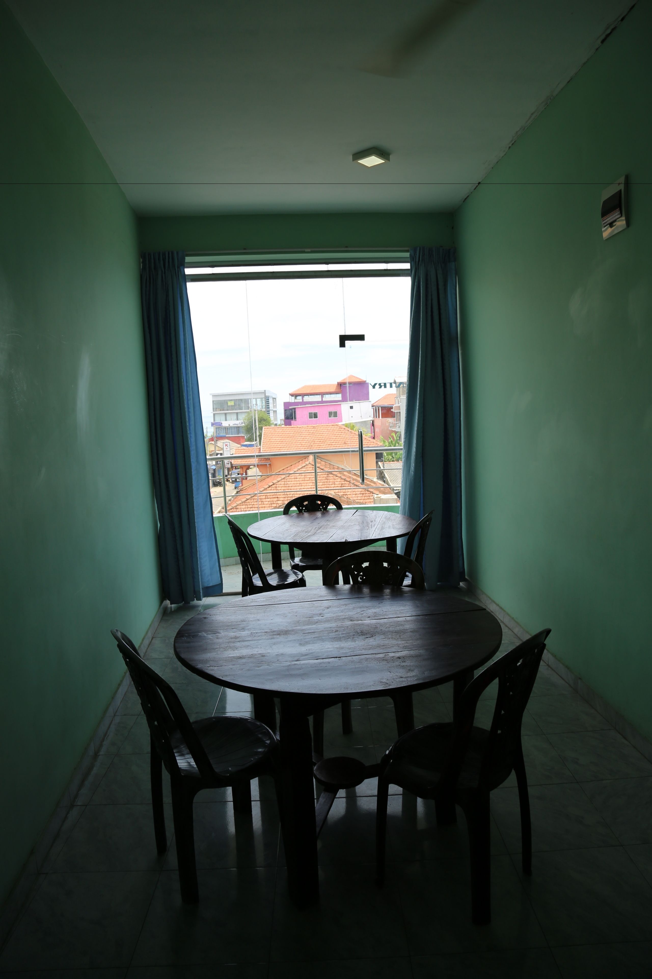 A room with a round table and four chairs, a window with blue curtains, and a view of buildings outside.