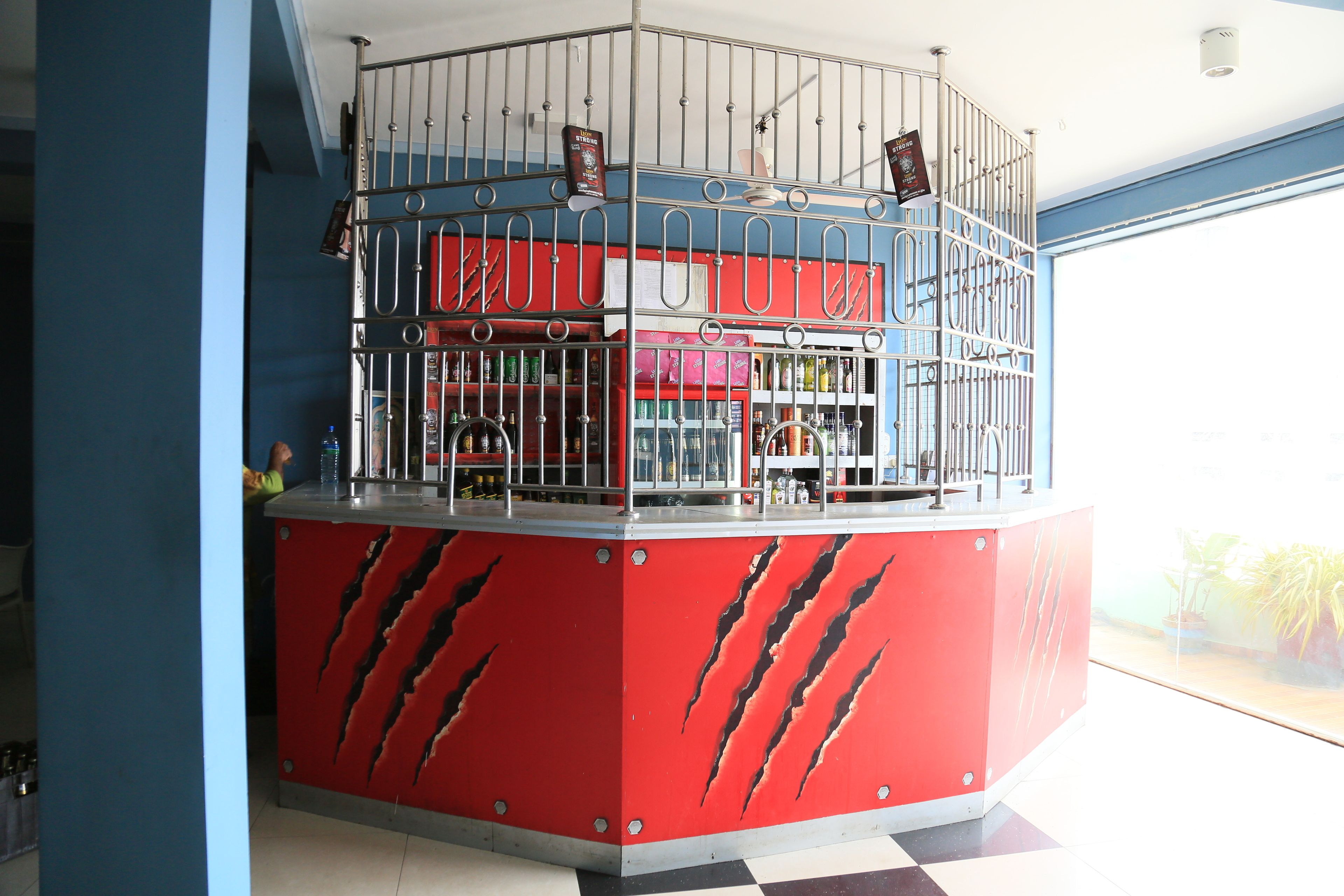 Un mostrador rojo con estructura de metal tipo jaula, estantes con botellas y una persona al fondo.