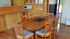 A wooden dining table surrounded by six chairs with cushioned seats, a white microwave on a wooden cabinet, and a window with a view of greenery. - Freycinet Sands (Coles Bay)