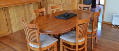Uma mesa de jantar de madeira cercada por seis cadeiras com assentos acolchoados, um micro-ondas branco sobre um armário de madeira e uma janela com vista para o verde.