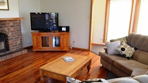 A living room with a brick fireplace, a television on a wooden cabinet, a wooden coffee table, and a beige sofa with patterned cushions. - Freycinet Sands (Coles Bay)